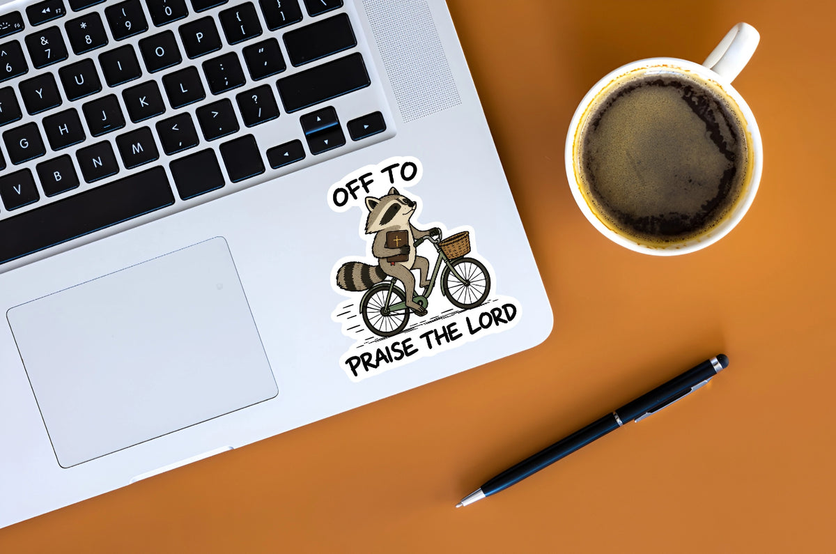 Off to Praise the Lord Christian raccoon sticker displayed on a silver laptop beside coffee
