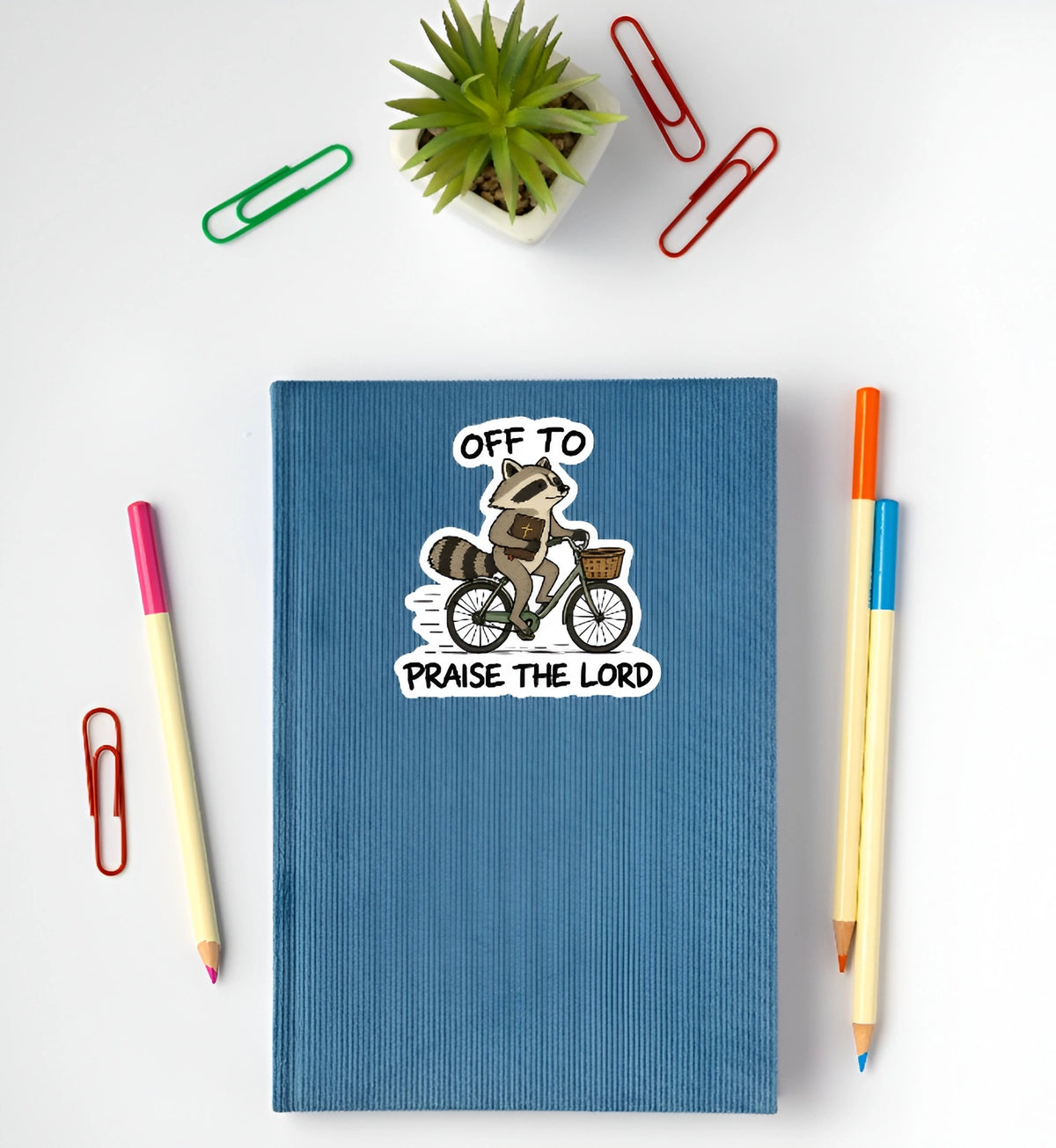 Cute Christian raccoon riding a bicycle with Bible sticker displayed on a blue notebook