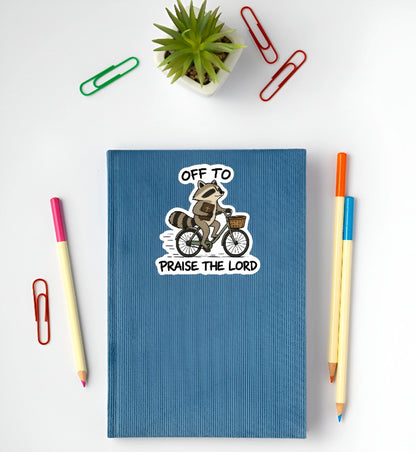 Cute Christian raccoon riding a bicycle with Bible sticker displayed on a blue notebook