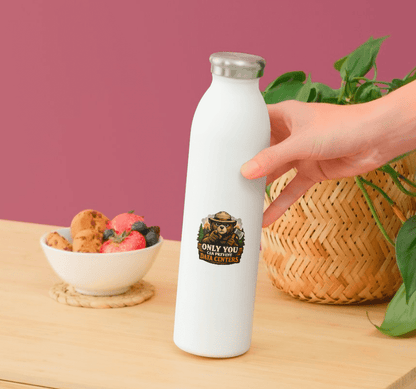 White water bottle with a logo held by a hand on a wooden table with a bowl of fruit and a plant in the background.