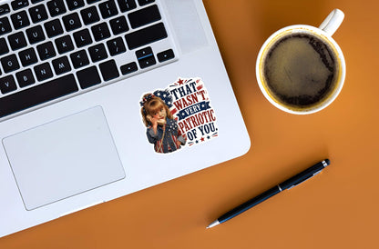 Laptop with a sticker featuring a detailed cartoon illustration of a sassy little girl holding a retro telephone receiver next to a distressed American flag background and bold red and blue typographic text reading "THAT WASN'T VERY PATRIOTIC OF YOU."on a desk next to a cup of coffee and pen