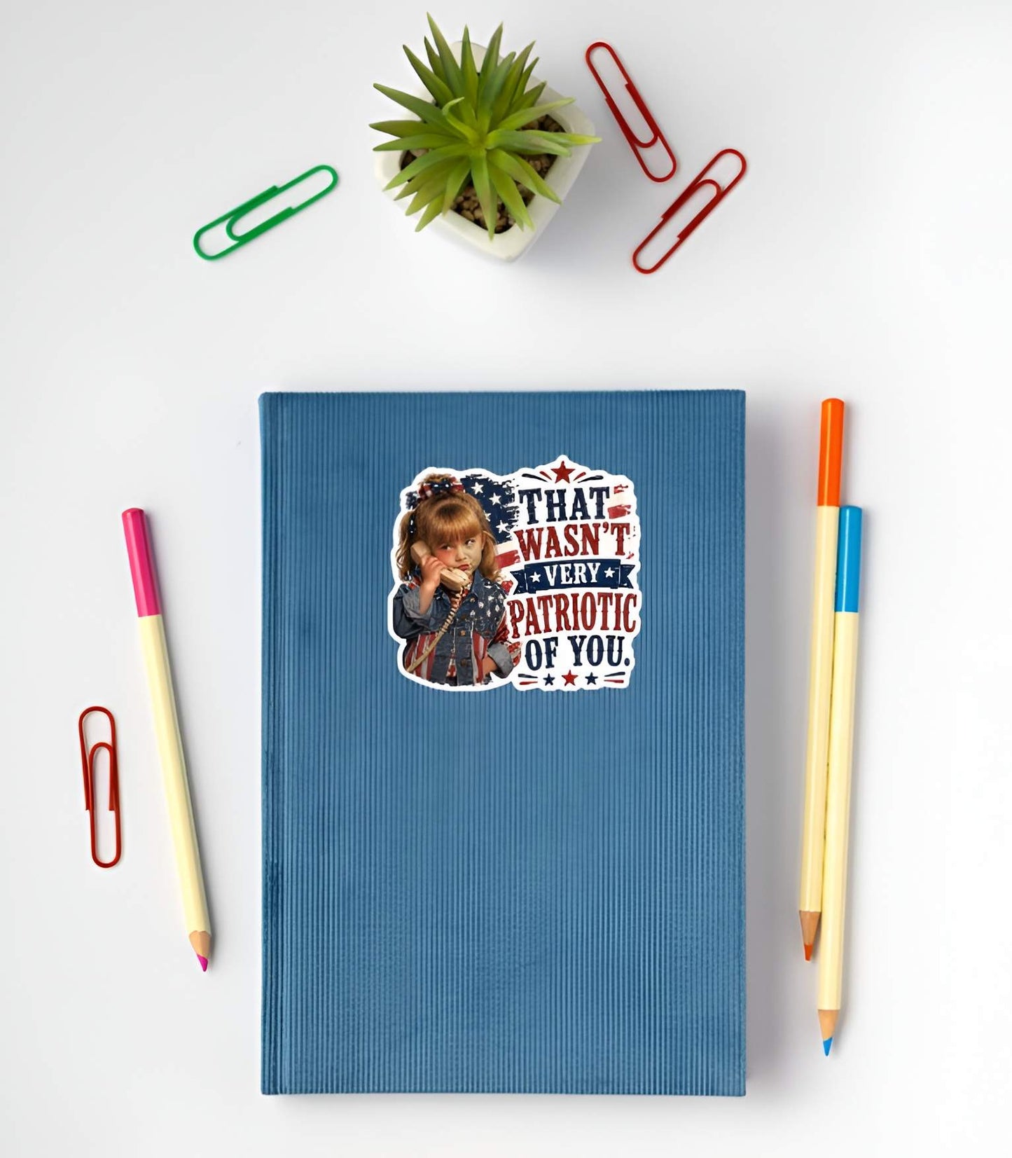 Blue notebook with a sticker featuring a detailed cartoon illustration of a sassy little girl holding a retro telephone receiver next to a distressed American flag background and bold red and blue typographic text reading "THAT WASN'T VERY PATRIOTIC OF YOU." on a white surface with stationery items