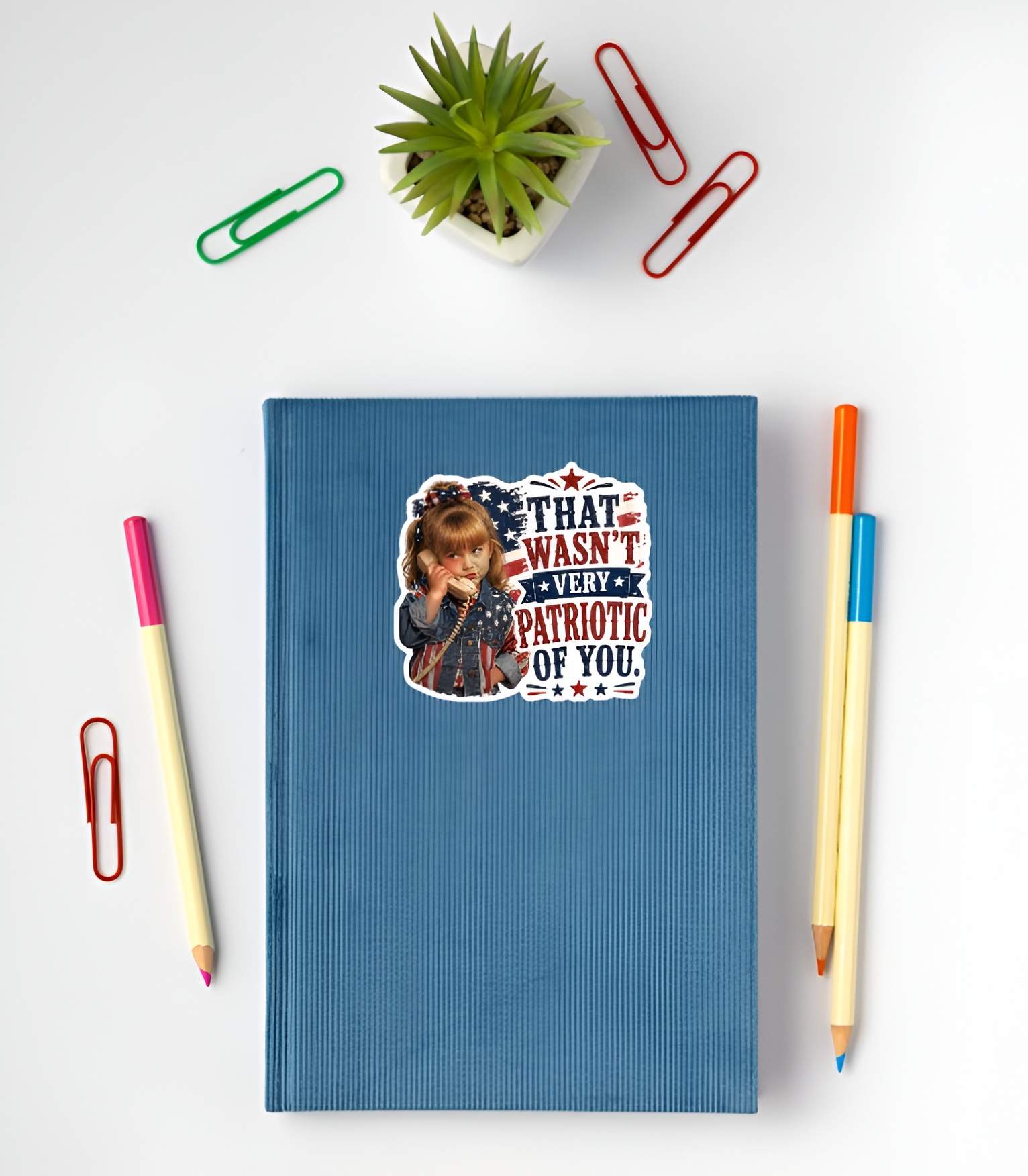 Blue notebook with a sticker featuring a detailed cartoon illustration of a sassy little girl holding a retro telephone receiver next to a distressed American flag background and bold red and blue typographic text reading "THAT WASN'T VERY PATRIOTIC OF YOU." on a white surface with stationery items