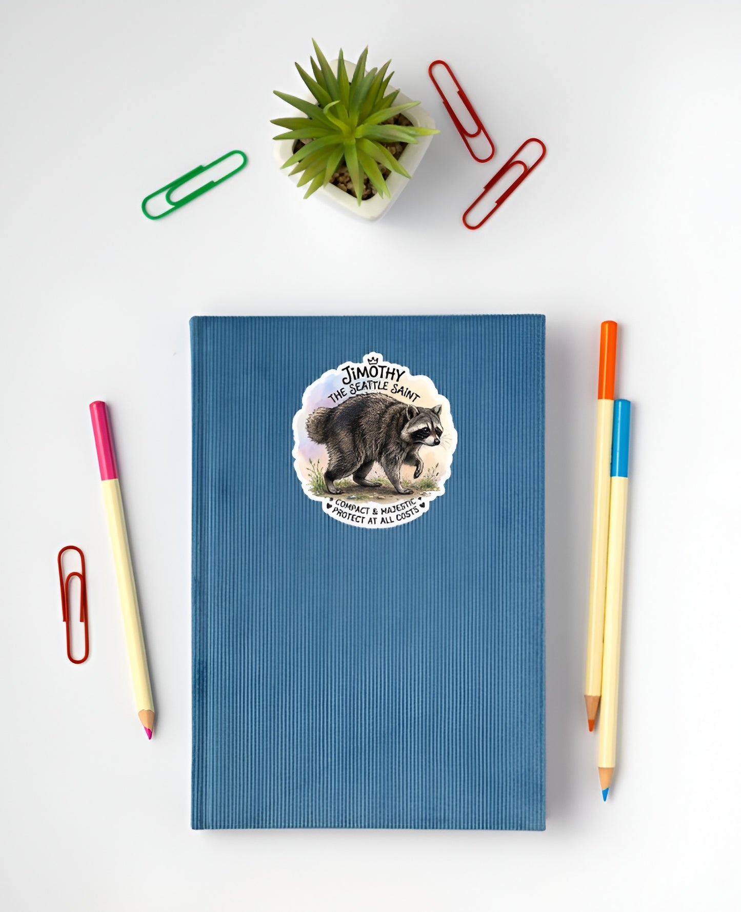A blue ribbed notebook or journal with a round raccoon illustration sticker on the front, arranged in a flat lay on a white background with paper clips, pencils, and a small potted succulent.