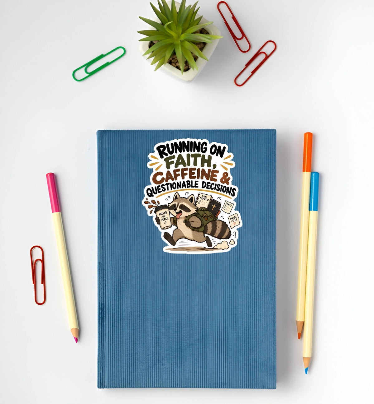A blue textured hardcover notebook with a cartoon graphic and the text 'RUNNING ON FAITH, CAFFEINE & QUESTIONABLE DECISIONS' lies on a white desk surrounded by stationery props including paperclips, pencils, and a small potted succulent.