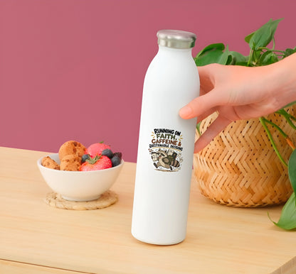 A hand holding a white insulated water bottle with a silver cap, featuring a graphic that reads 'RUNNING ON FAITH, CAFFEINE & GENTLE REMEDIES.' The bottle is set on a wooden table with a bowl of fruit, a woven basket, and green leaves against a pink background.