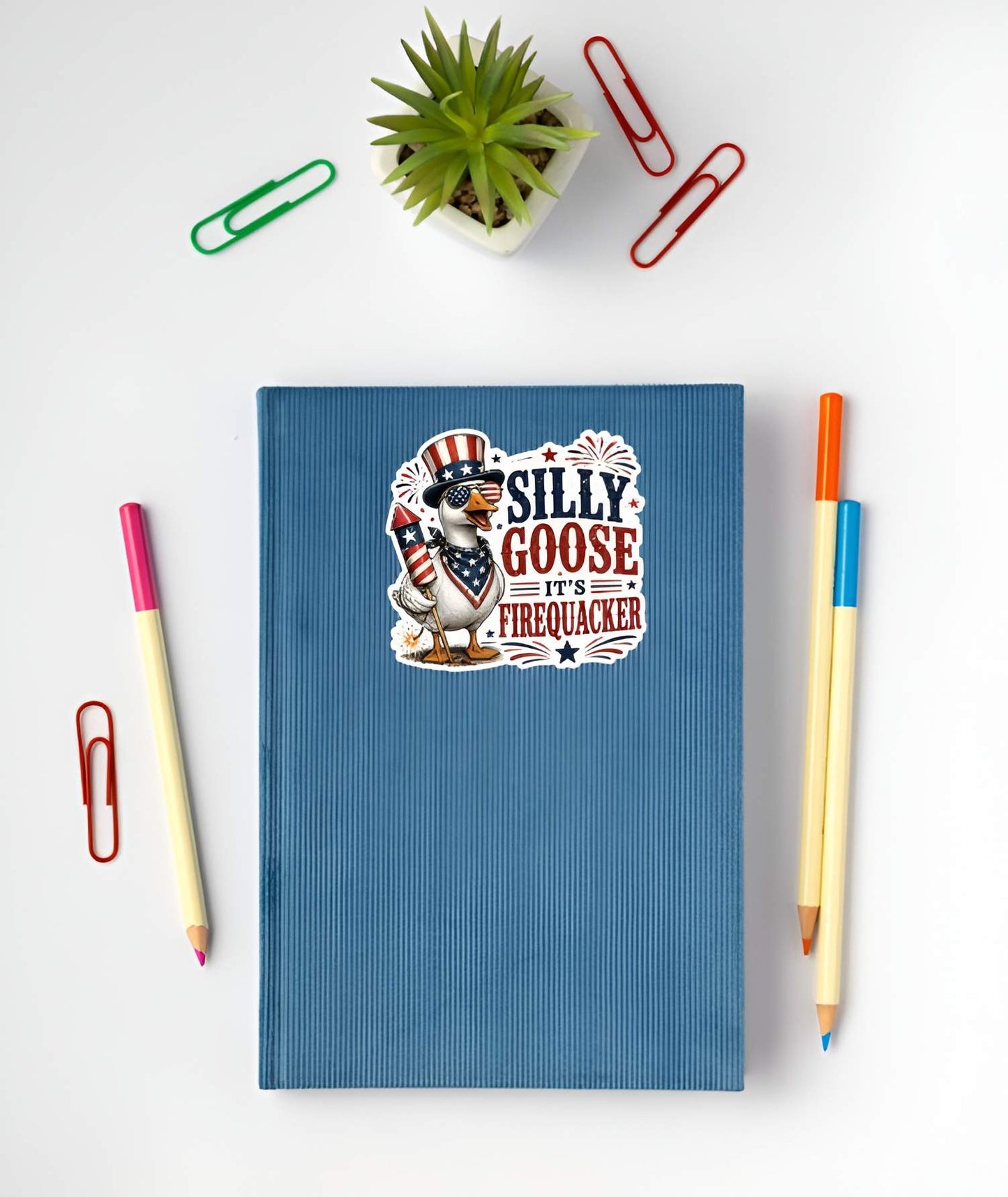 Blue notebook with a patriotic silly goose sticker on a white surface with stationery items.