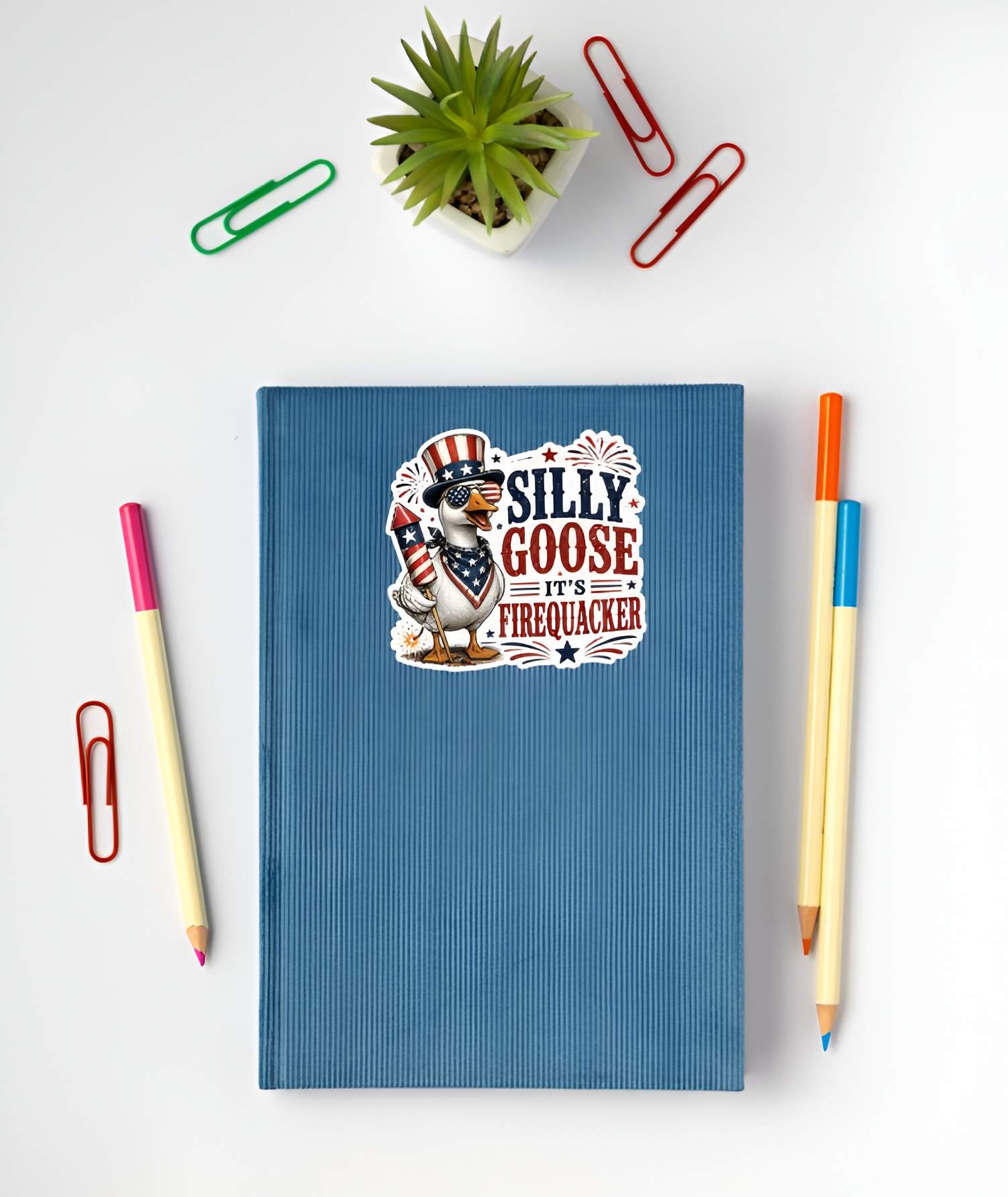 Blue notebook with a patriotic silly goose sticker on a white surface with stationery items.