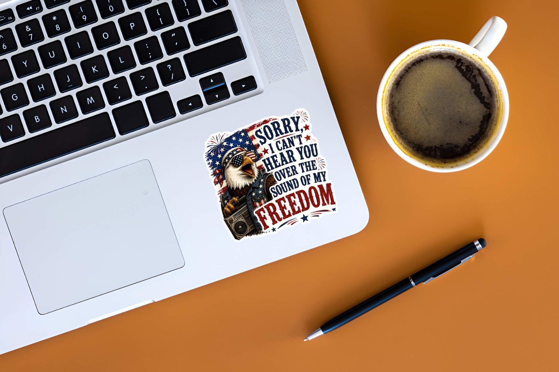 Laptop with a sticker featuring a detailed cartoon illustration of a squawking bald eagle wearing flag sunglasses, a bandana, and a denim vest while holding a retro boombox radio, next to bold text reading "SORRY, I CAN'T HEAR YOU OVER THE SOUND OF MY FREEDOM", coffee cup, and pen on an orange surface
