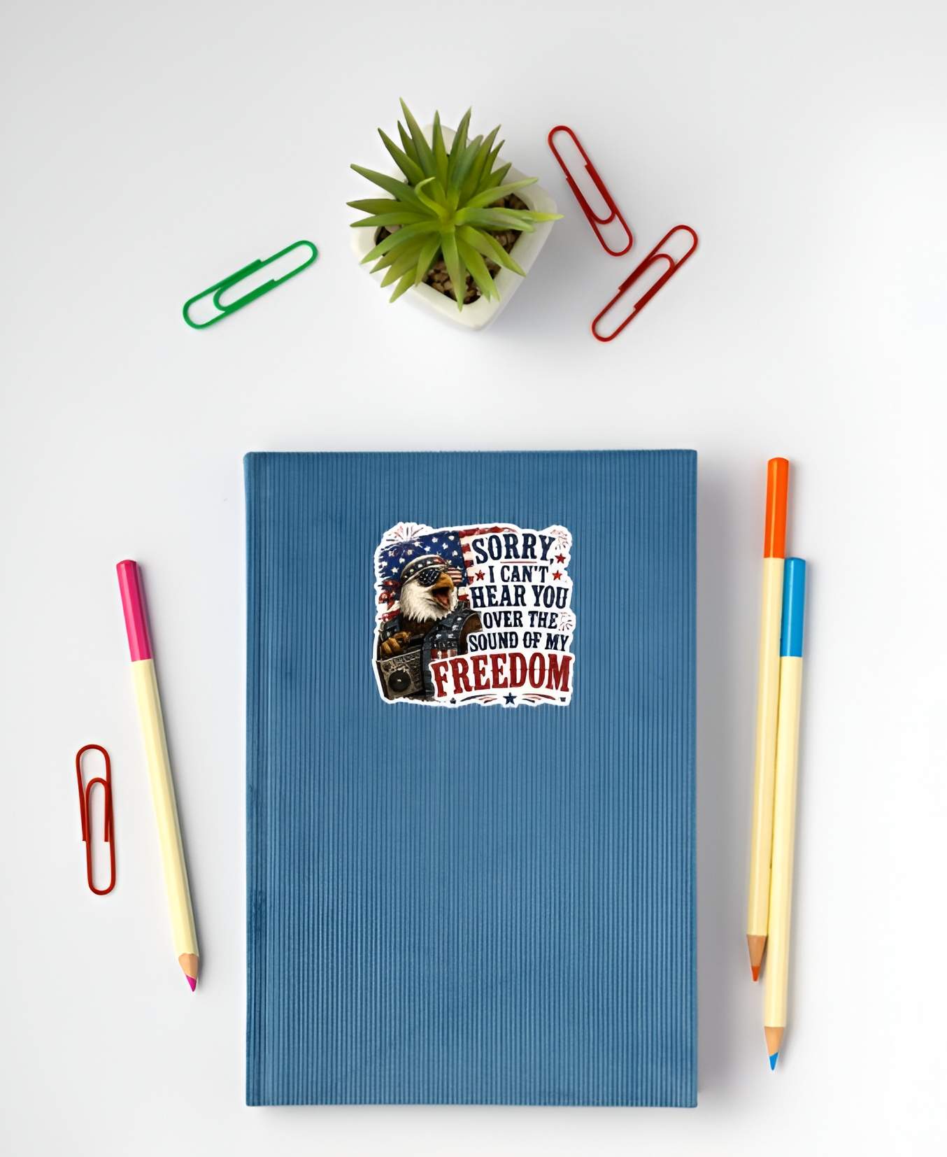 Blue notebook with a sticker featuring a detailed cartoon illustration of a squawking bald eagle wearing flag sunglasses, a bandana, and a denim vest while holding a retro boombox radio, next to bold text reading "SORRY, I CAN'T HEAR YOU OVER THE SOUND OF MY FREEDOM".on a white surface with stationery items