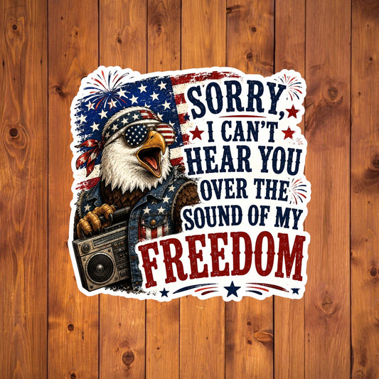 Sticker with an eagle holding a boombox and text 'Sorry, I can't hear you over the sound of my freedom' on a wooden background.