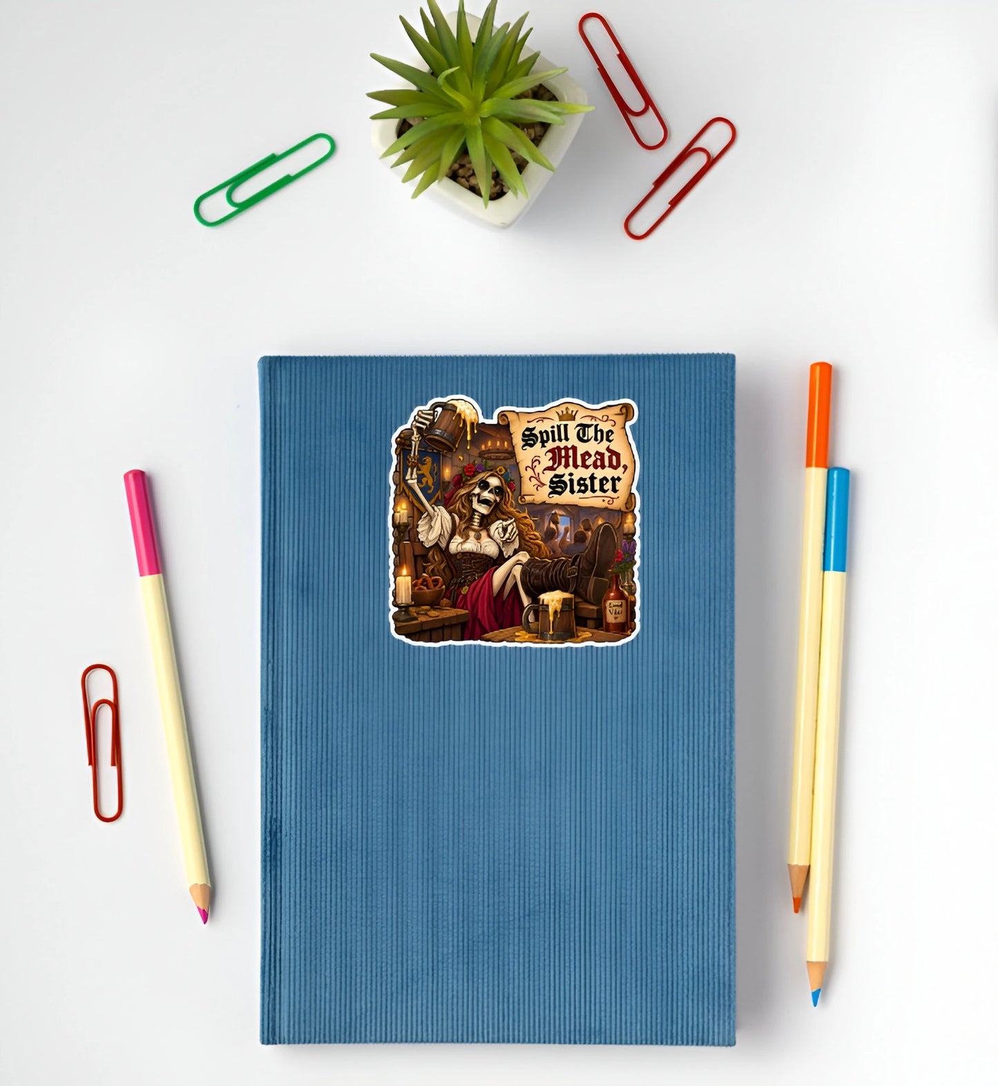 A blue ribbed hardcover notebook with a die-cut sticker of a skeleton serving wine and the text 'Spill The Mead Sister' on the front. The notebook is styled in a flat-lay arrangement on a white background, surrounded by colorful paperclips, pencils, and a small potted succulent.