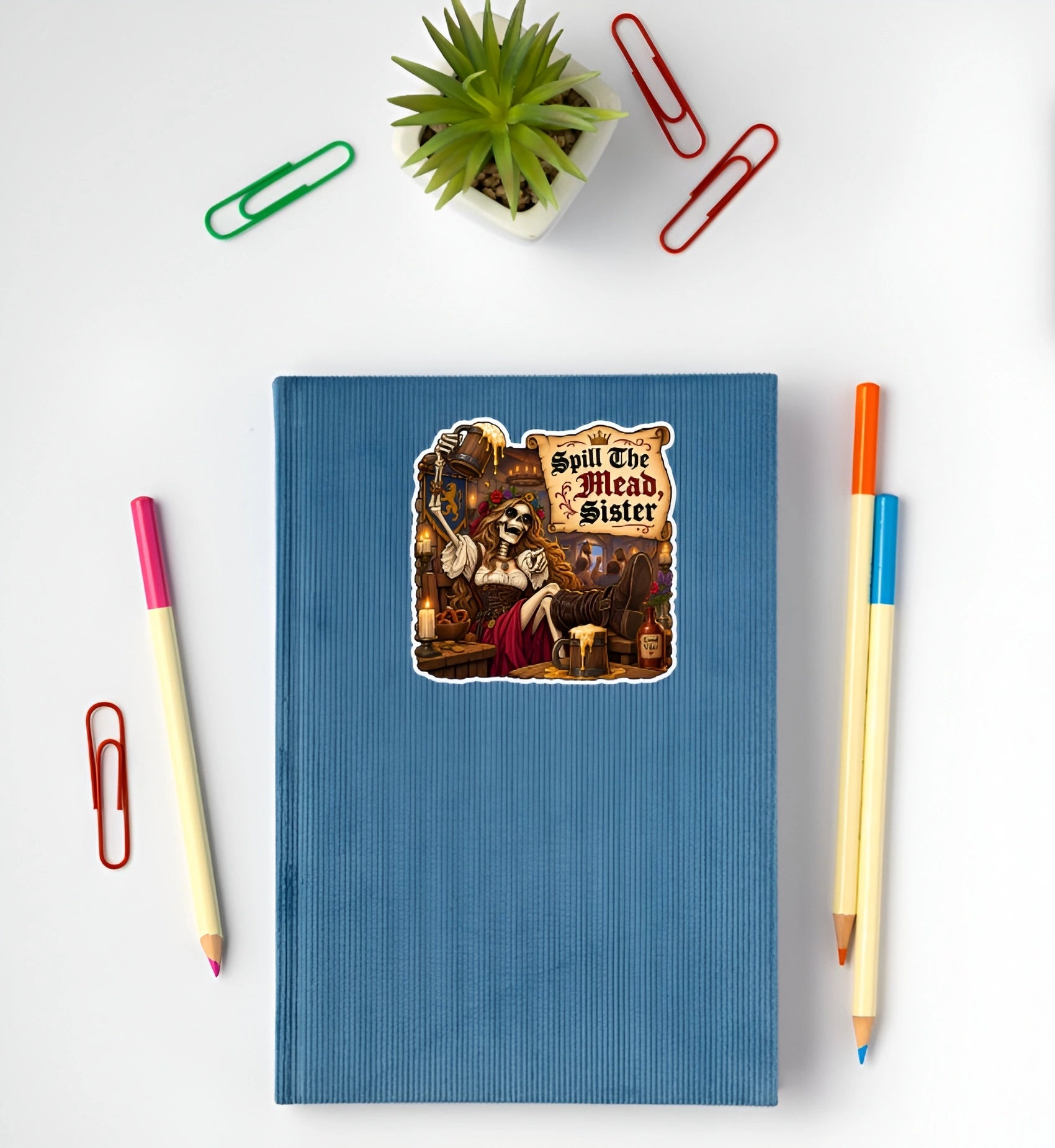 A blue ribbed hardcover notebook with a die-cut sticker of a skeleton serving wine and the text 'Spill The Mead Sister' on the front. The notebook is styled in a flat-lay arrangement on a white background, surrounded by colorful paperclips, pencils, and a small potted succulent.