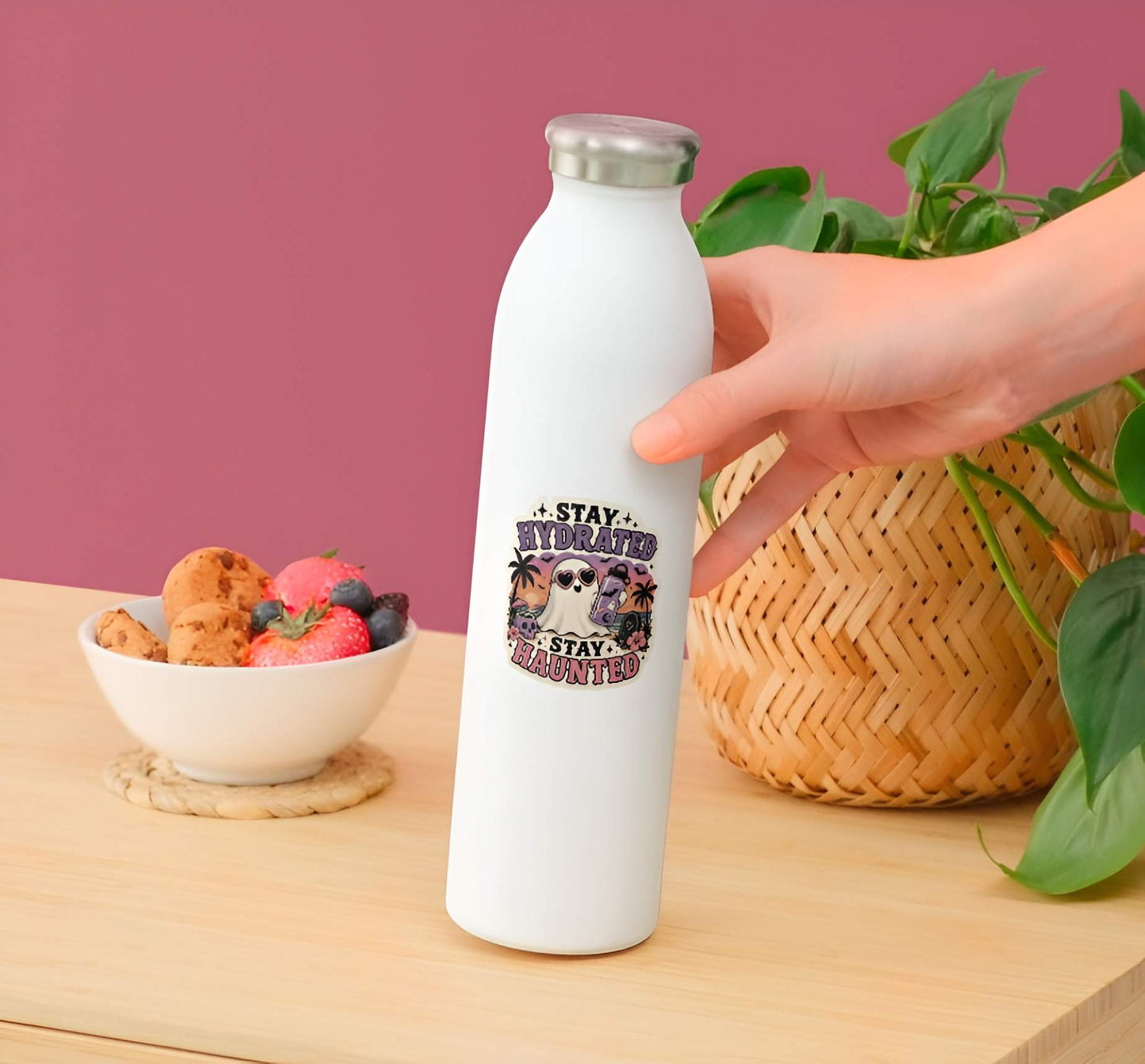 White water bottle with a tropical ghost with purple flask and heart sunglasses, sunset beach skull drink graphic. on a wooden table with a pink background