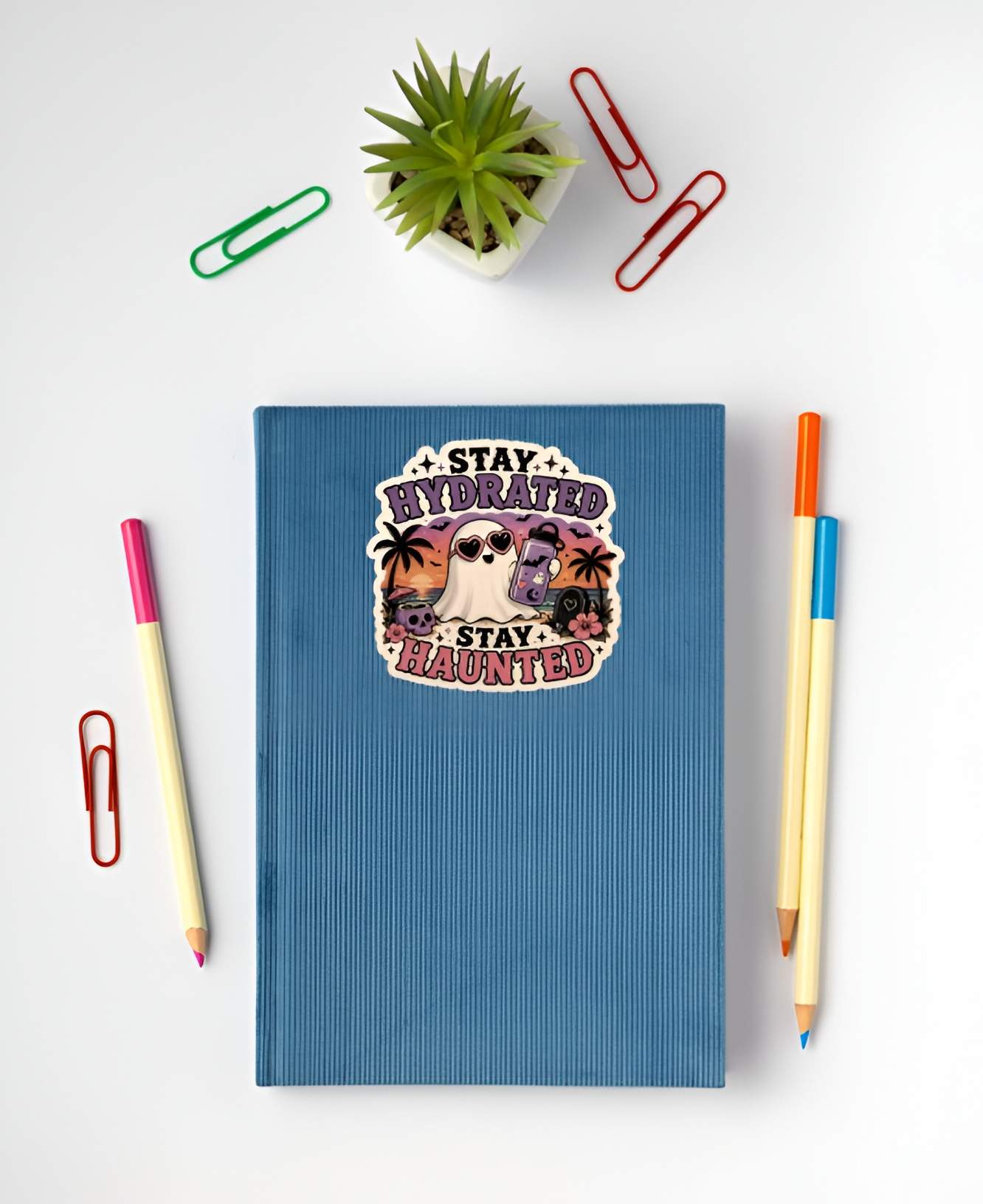 Blue notebook with a tropical ghost with purple flask and heart sunglasses, sunset beach skull drink graphic. and text on a white background with stationery items.