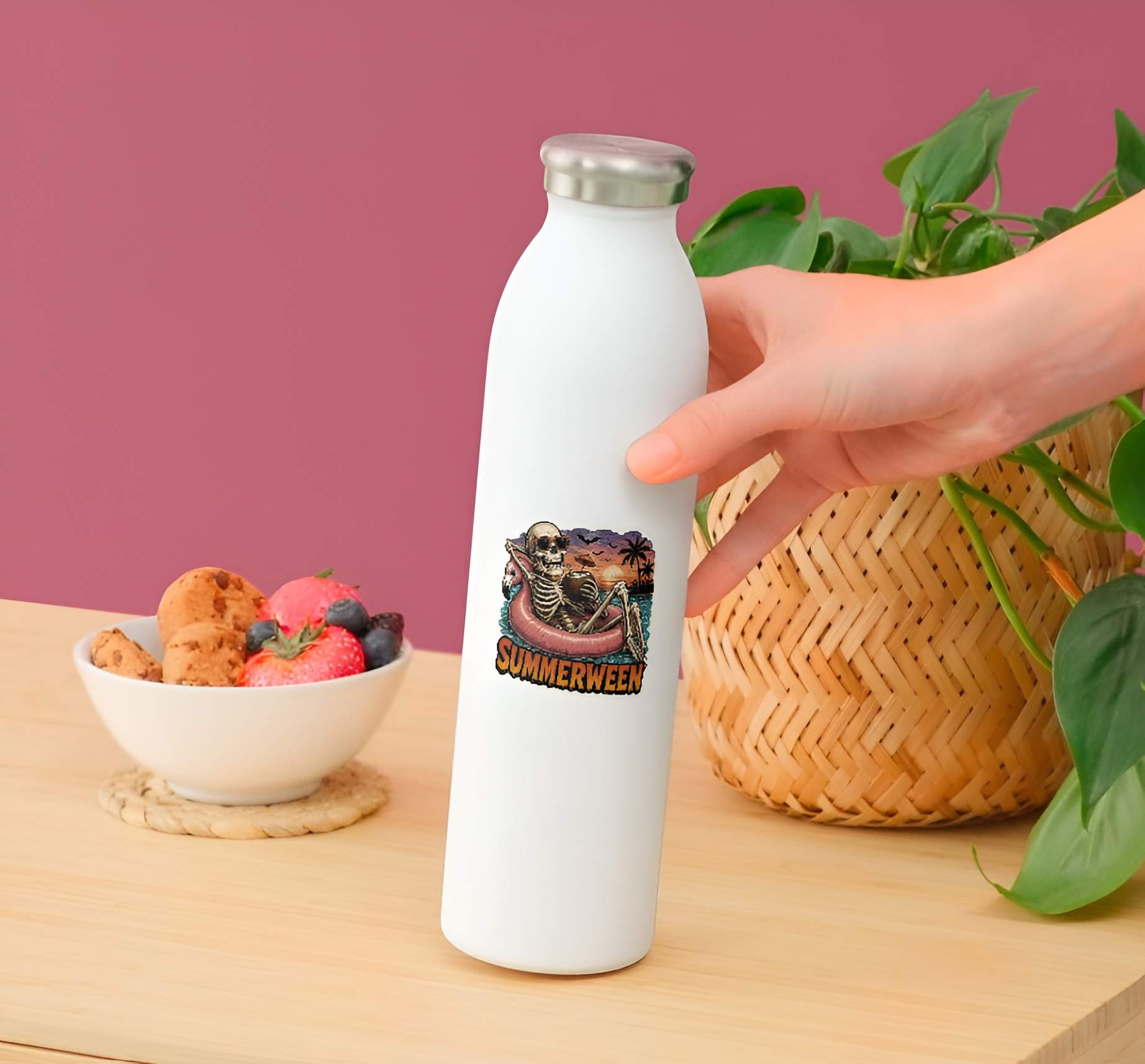 White water bottle with a design of a Funny Summerween skeleton sticker on a wooden table with a bowl of snacks and a plant.
