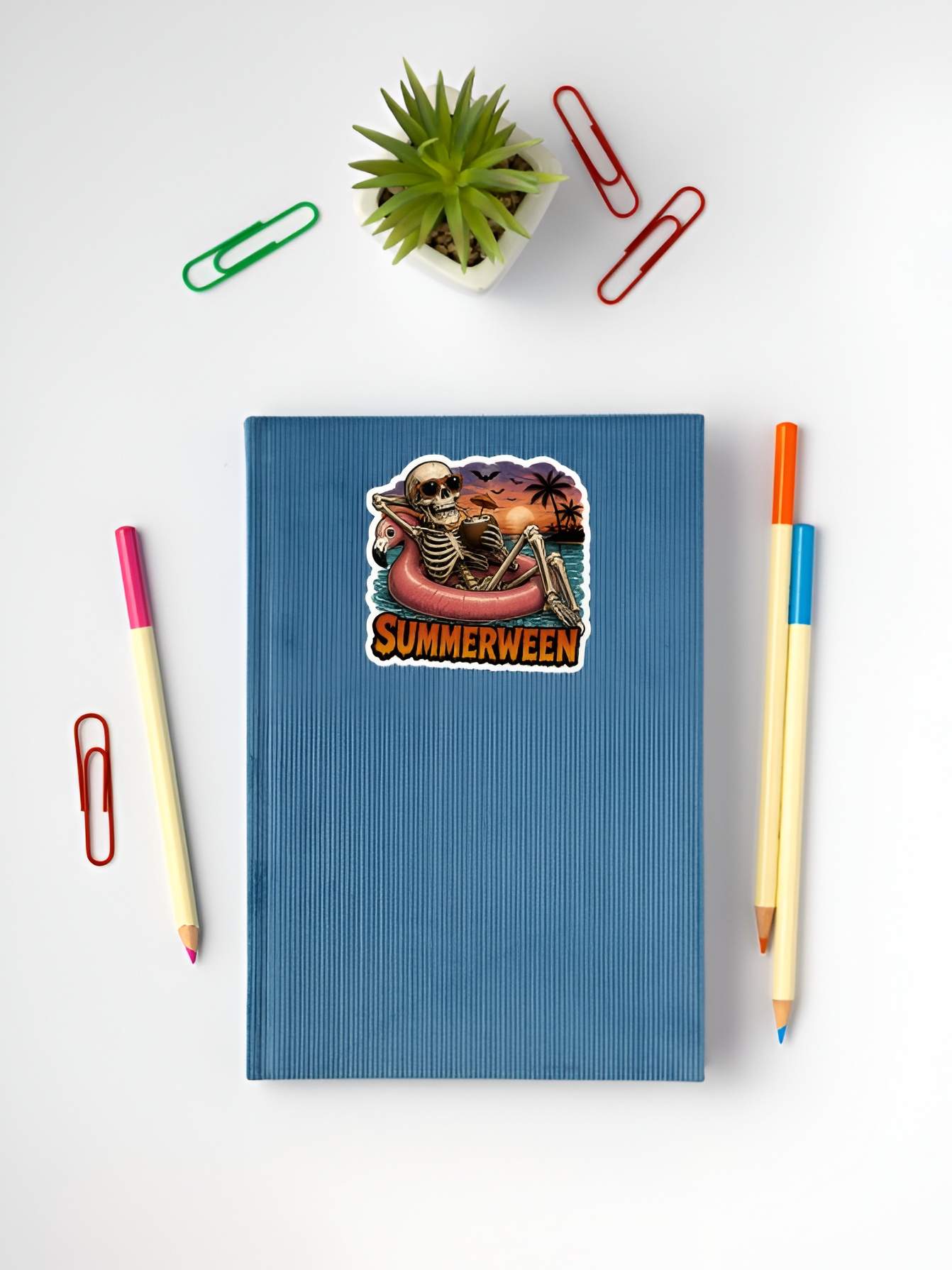 Blue notebook with a 'Summerween' skeleton sticker, surrounded by stationery items on a white background.