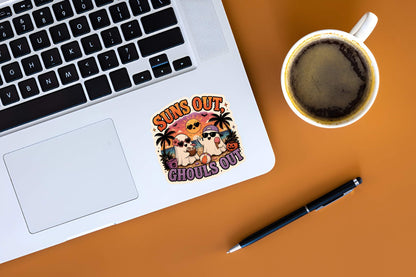Laptop with a Funny Suns Out Ghouls Out ghost sticker on a desk next to a cup of coffee and pen