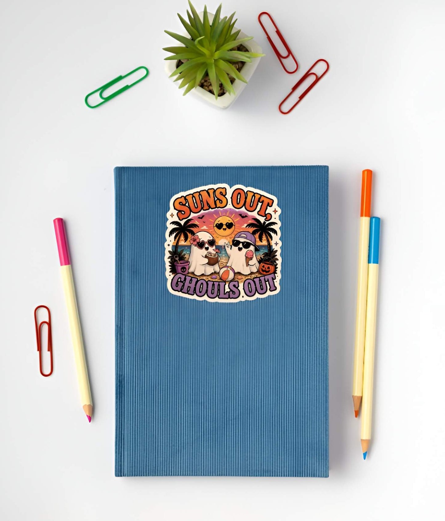 Blue notebook with a Funny Suns Out Ghouls Out ghost sticker on the cover, surrounded by stationery items on a white background.