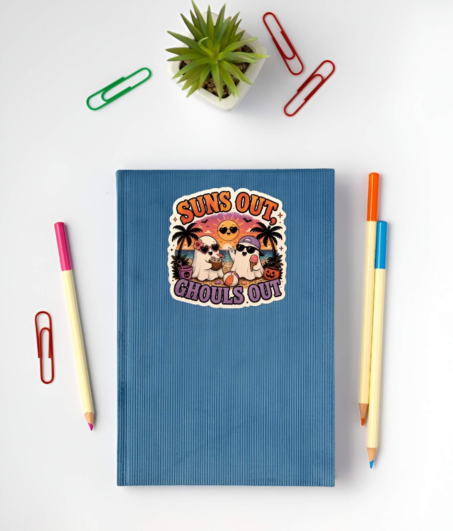 Blue notebook with a Funny Suns Out Ghouls Out ghost sticker on the cover, surrounded by stationery items on a white background.