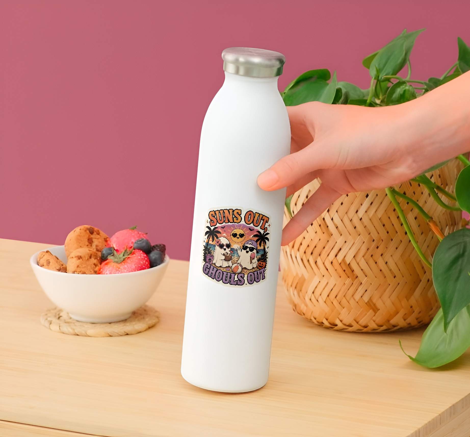 White water bottle with a Funny Suns Out Ghouls Out ghost sticker held by a hand on a wooden surface with a pink background.