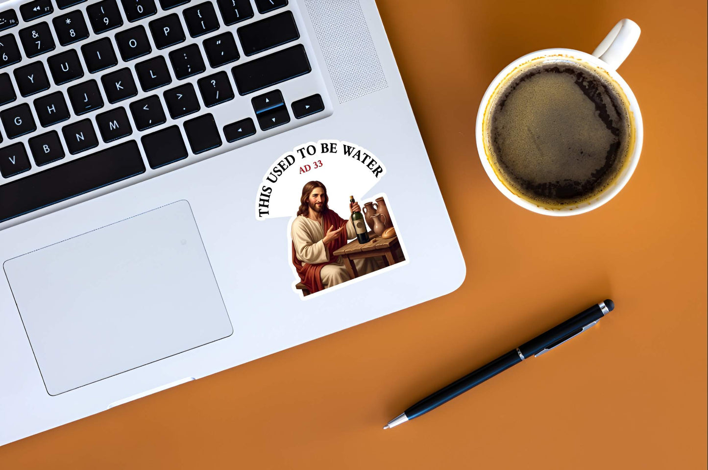Laptop with a sticker of Jesus sitting at a wooden table, smiling and holding a corked wine bottle next to clay jugs and bread, underneath an arched text box reading "THIS USED TO BE WATER" and a cup of coffee on a brown surface