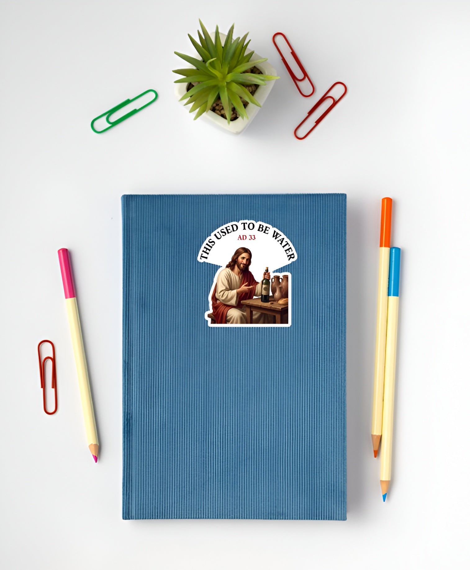 Blue notebook with a sticker  of Jesus sitting at a wooden table, smiling and holding a corked wine bottle next to clay jugs and bread, underneath an arched text box reading "THIS USED TO BE WATER" on a white surface with stationery items.