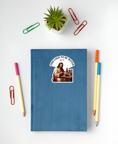 Blue notebook with a sticker  of Jesus sitting at a wooden table, smiling and holding a corked wine bottle next to clay jugs and bread, underneath an arched text box reading "THIS USED TO BE WATER" on a white surface with stationery items.