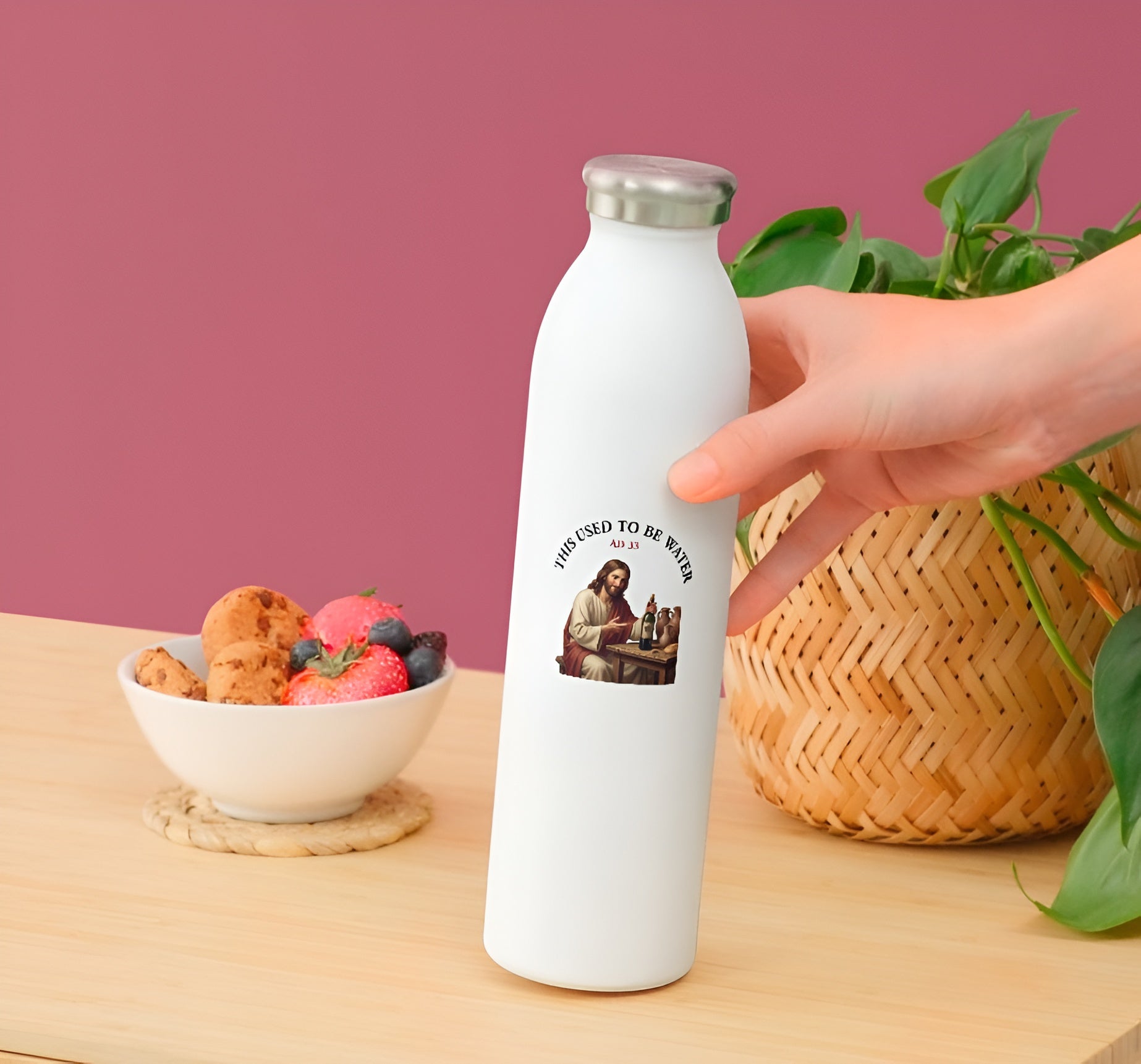 White water bottle with illustration of Jesus sitting at a wooden table, smiling and holding a corked wine bottle next to clay jugs and bread, underneath an arched text box reading "THIS USED TO BE WATER"held by a hand on a wooden table with a bowl of fruit and waffles.
