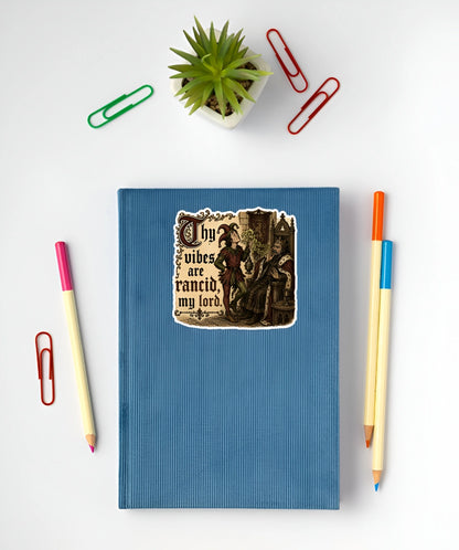 Thy Vibes Are Rancid My Lord medieval meme sticker displayed on a blue notebook