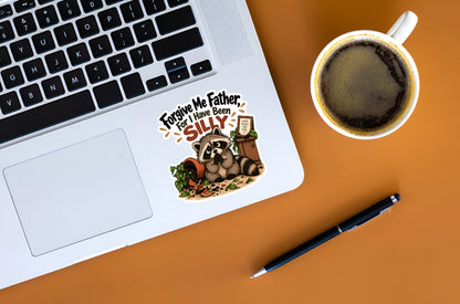 A die-cut sticker featuring a raccoon and the text 'Forgive Me Father, For I Have Been SILLY' is shown on the palm rest of a laptop. The laptop is placed on an orange desk alongside a cup of coffee and a pen.