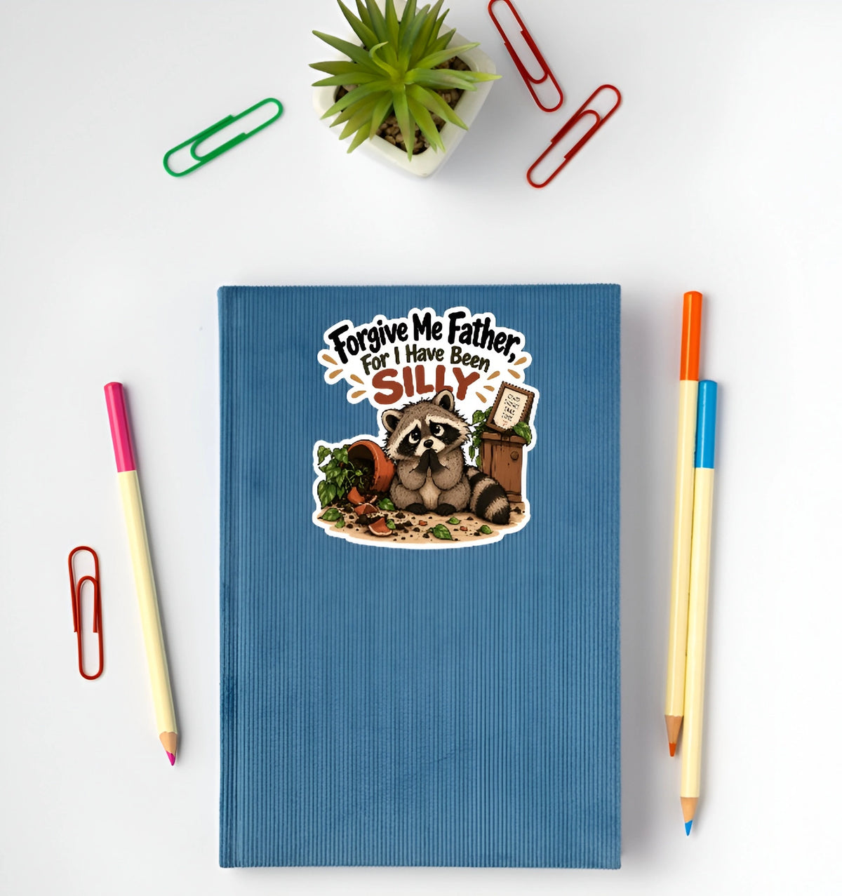 A blue ribbed hardcover notebook with a cartoon raccoon sticker and the text 'Forgive Me Father, For I Have Been SILLY' is shown in a flat-lay arrangement on a white background, surrounded by colored paperclips, pencils, and a small potted succulent.