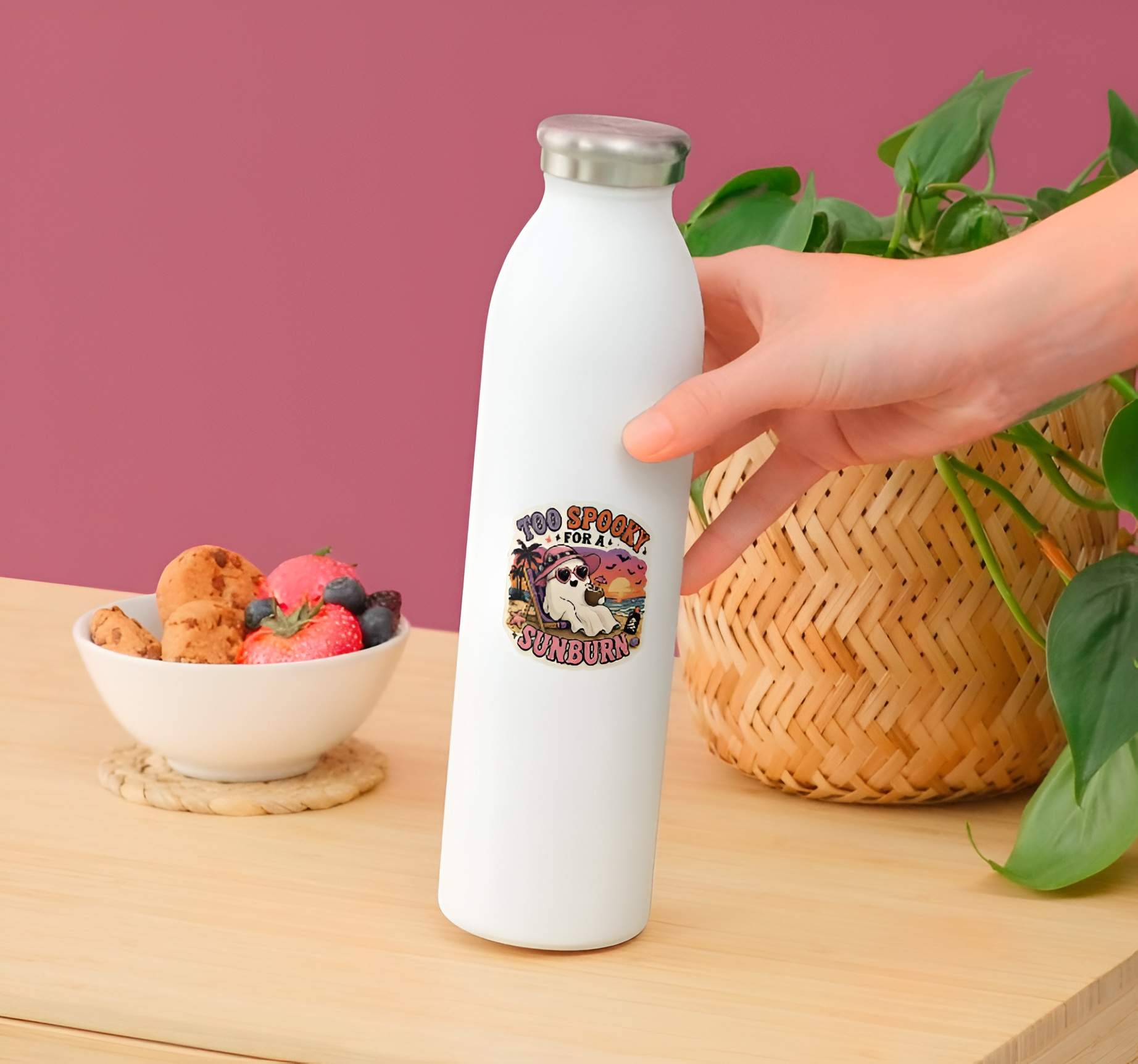 White water bottle with a Funny Too Spooky For A Sunburn ghost sticker, cute Halloween beach vinyl decal, tropical ghost sunbathing in pink hat and heart sunglasses, sunset coconut drink graphic. on a wooden table with a bowl of fruit and a plant.