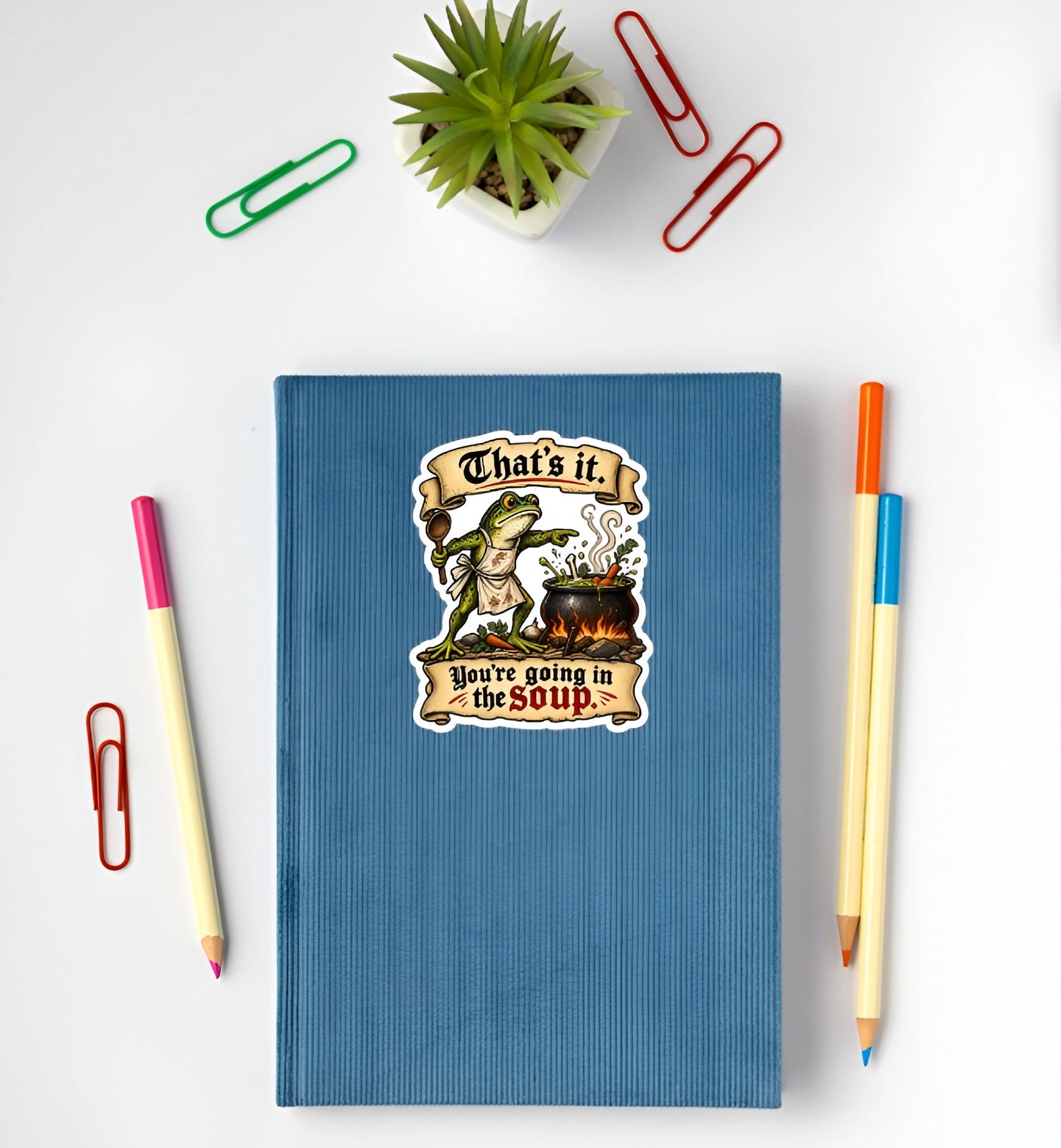 Funny medieval frog You’re Going in the Soup sticker displayed on a blue notebook
