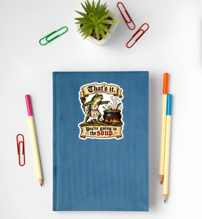 Funny medieval frog You’re Going in the Soup sticker displayed on a blue notebook