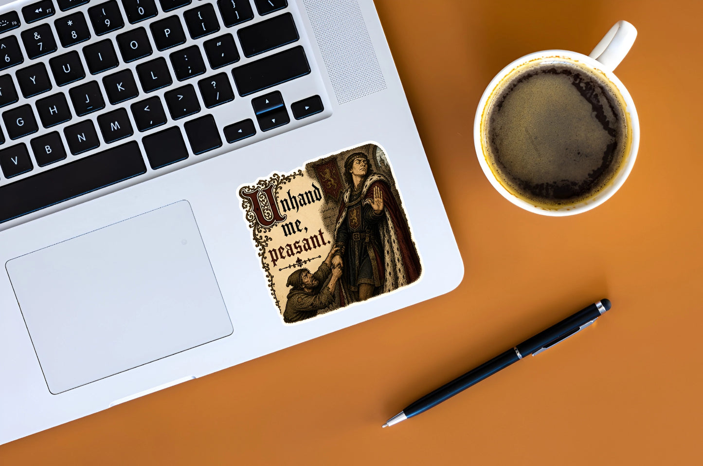 Unhand Me Peasant medieval meme sticker displayed on a silver laptop beside coffee