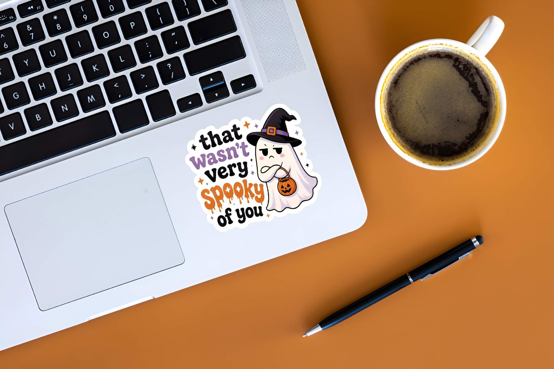 Laptop with a judging little ghost wearing a witch hat with arms crossed and a pumpkin basket next to bold text reading "that wasn't very spooky of you" sticker, coffee cup, and pen on an orange surface