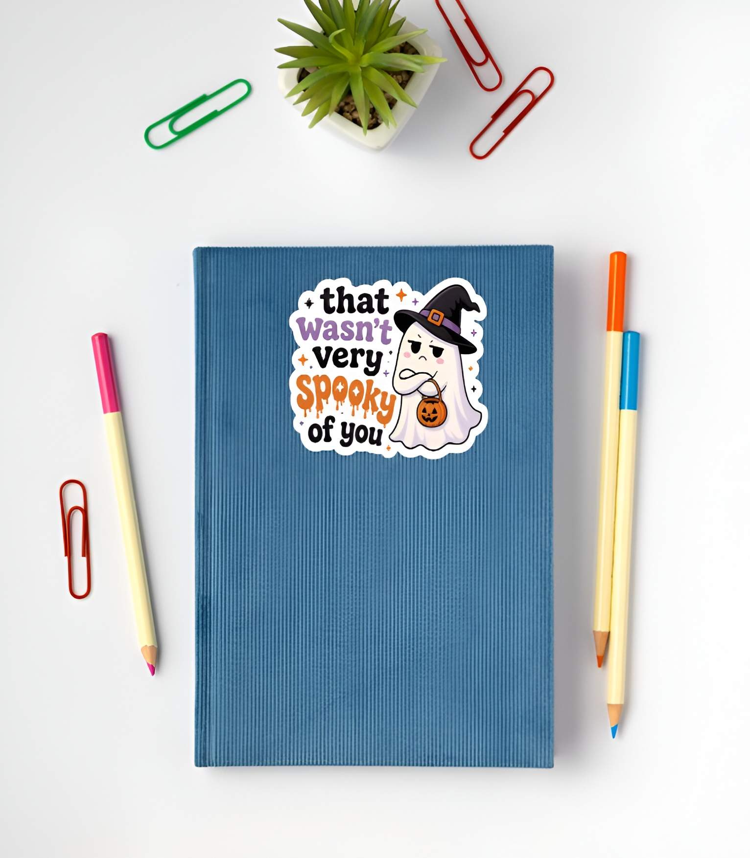 Blue notebook with a judging little ghost wearing a witch hat with arms crossed and a pumpkin basket next to bold text reading "that wasn't very spooky of you" sticker on a white surface with stationery items.