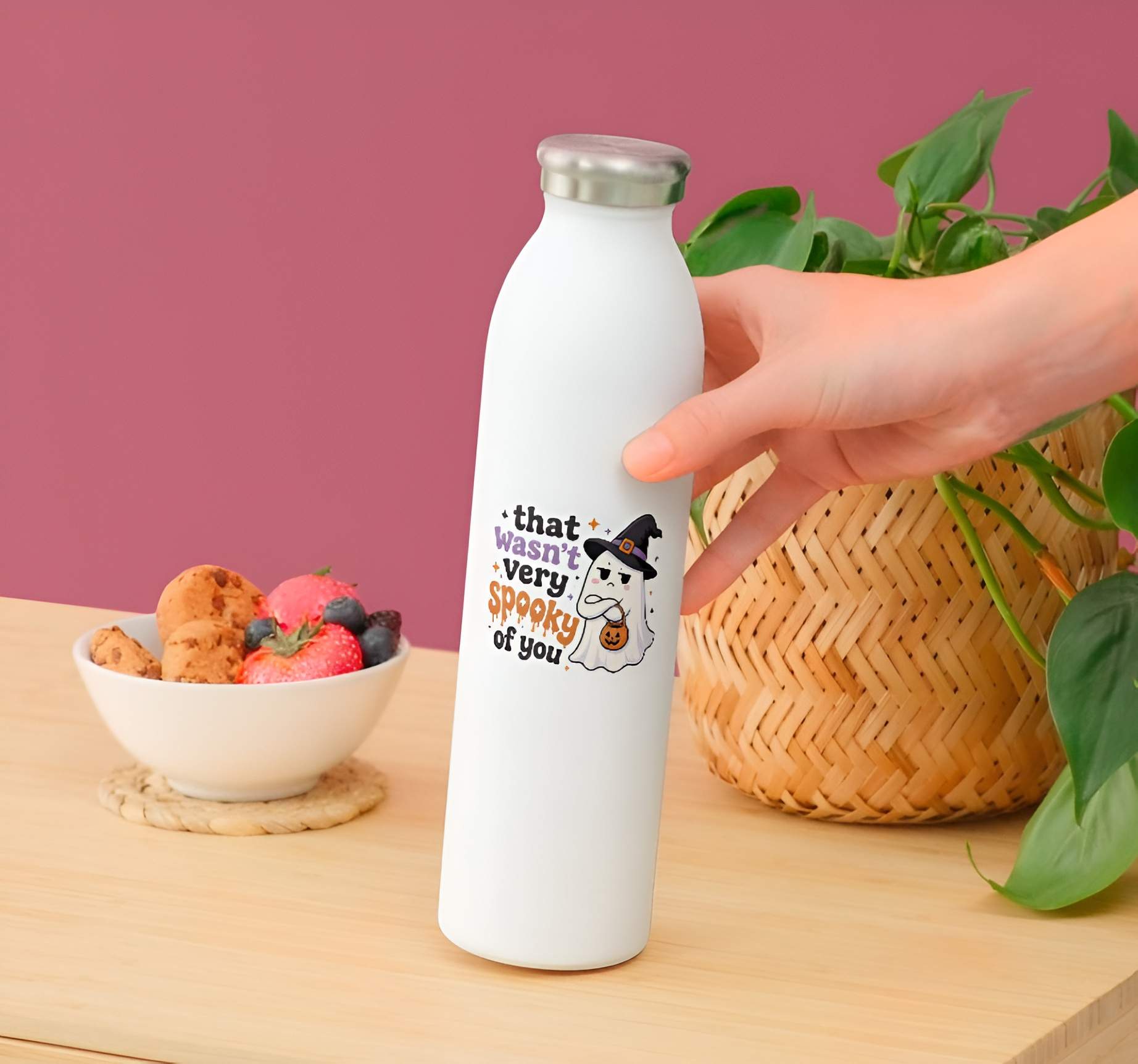 White water bottle with judging little ghost wearing a witch hat with arms crossed and a pumpkin basket next to bold text reading "that wasn't very spooky of you" by a hand on a wooden table with a bowl of snacks and plant in the background.