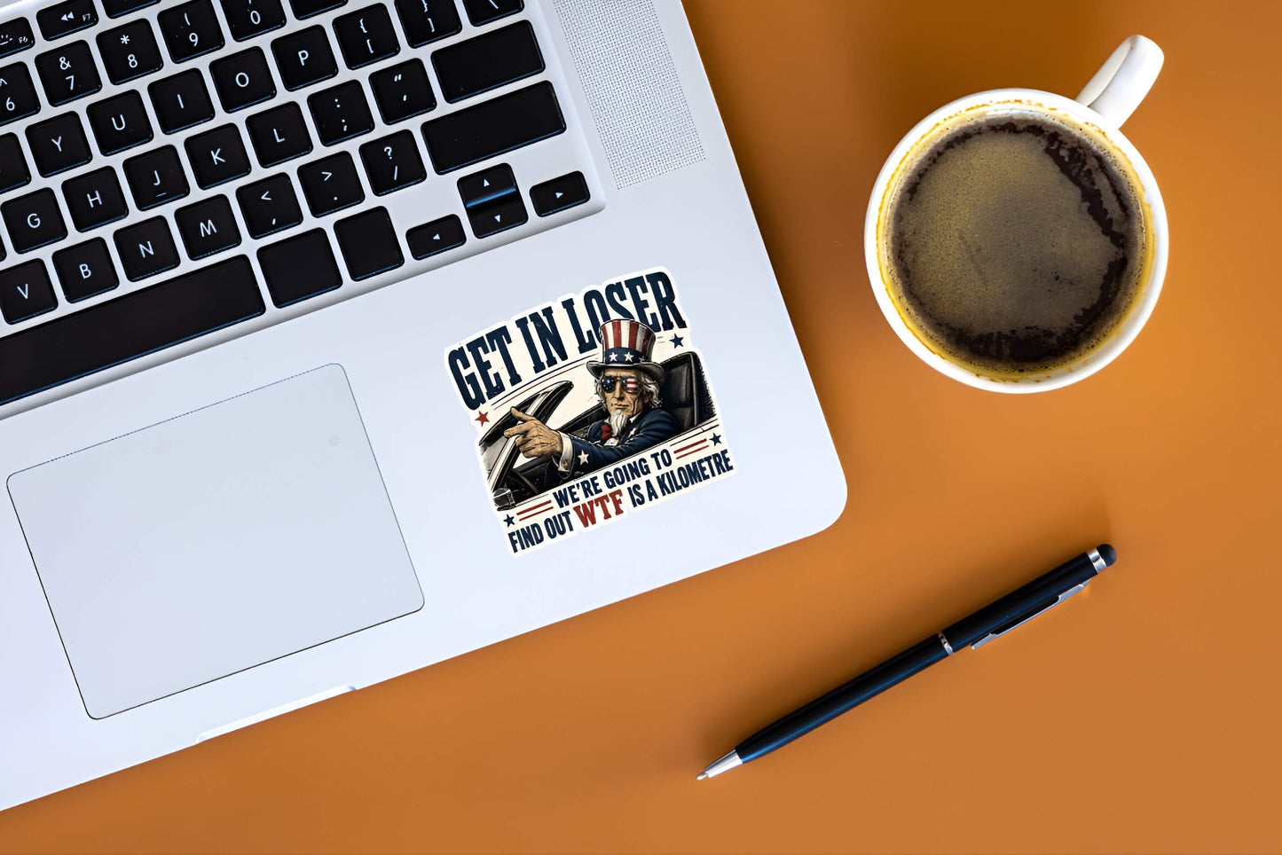 Laptop with a sticker sticker featuring a detailed vintage-style illustration of Uncle Sam wearing flag sunglasses and driving a convertible car, pointing forward next to bold text reading "GET IN LOSER WE'RE GOING TO FIND OUT WTF IS A KILOMETRE". on a desk next to a coffee cup and pen