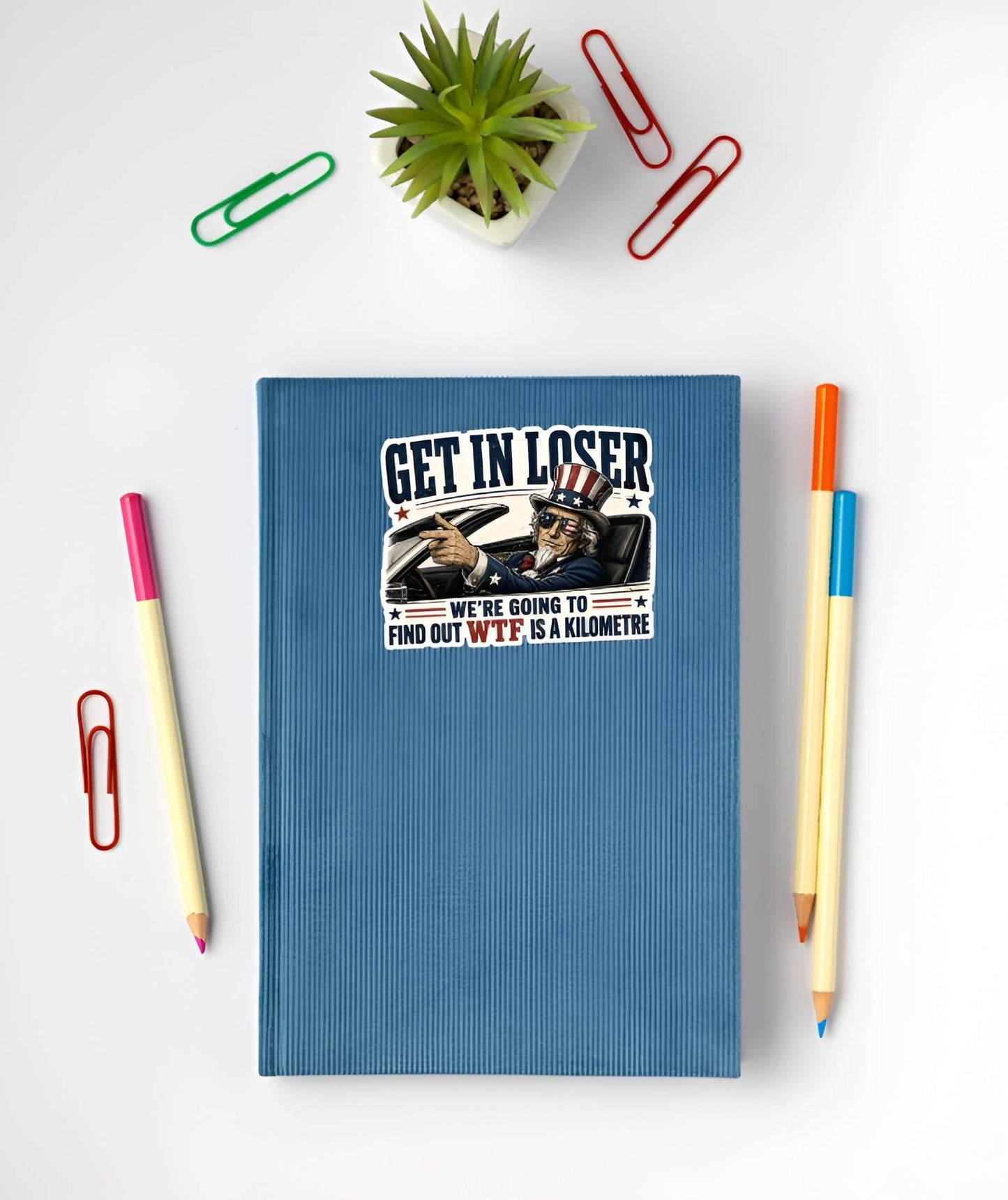 Blue notebook with a sticker featuring a detailed vintage-style illustration of Uncle Sam wearing flag sunglasses and driving a convertible car, pointing forward next to bold text reading "GET IN LOSER WE'RE GOING TO FIND OUT WTF IS A KILOMETRE". on a white surface with stationery items.