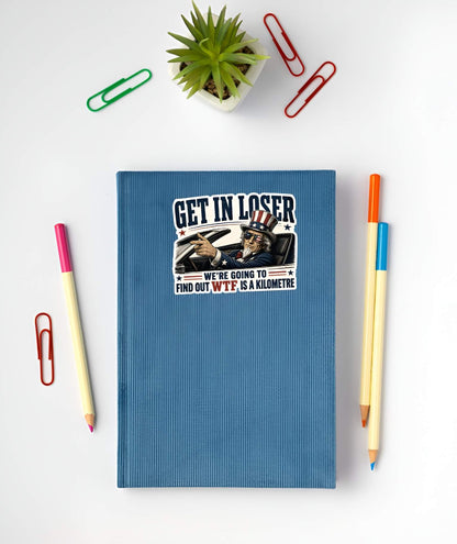 Blue notebook with a sticker featuring a detailed vintage-style illustration of Uncle Sam wearing flag sunglasses and driving a convertible car, pointing forward next to bold text reading "GET IN LOSER WE'RE GOING TO FIND OUT WTF IS A KILOMETRE". on a white surface with stationery items.