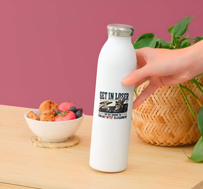 White water bottle with a sticker  featuring a detailed vintage-style illustration of Uncle Sam wearing flag sunglasses and driving a convertible car, pointing forward next to bold text reading "GET IN LOSER WE'RE GOING TO FIND OUT WTF IS A KILOMETRE". on a wooden table with a bowl of fruit and plant in the background.