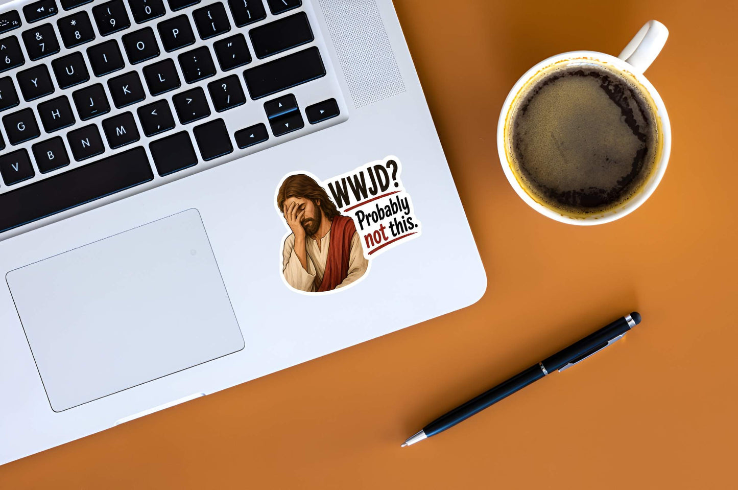 Laptop with a Jesus wearing cream and red robes, holding his hand over his face in an exhausted facepalm gesture next to bold text reading "WWJD? Probably not this." sticker on the palm rest area, next to a cup of coffee and a pen on a brown surface.