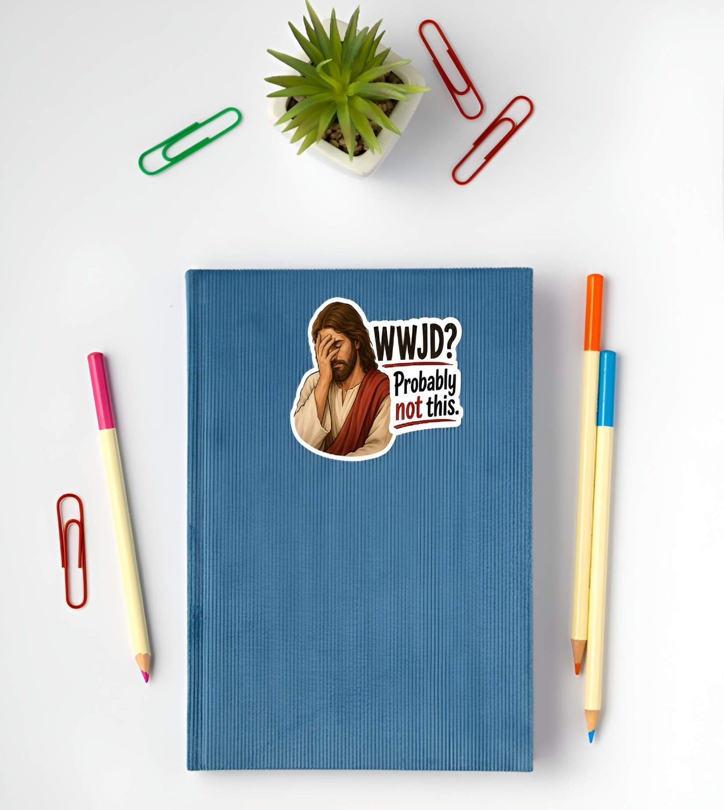 Blue notebook with a Jesus wearing cream and red robes, holding his hand over his face in an exhausted facepalm gesture next to bold text reading "WWJD? Probably not this."sticker of a cartoon character and text, surrounded by pens and paper clips on a white background.