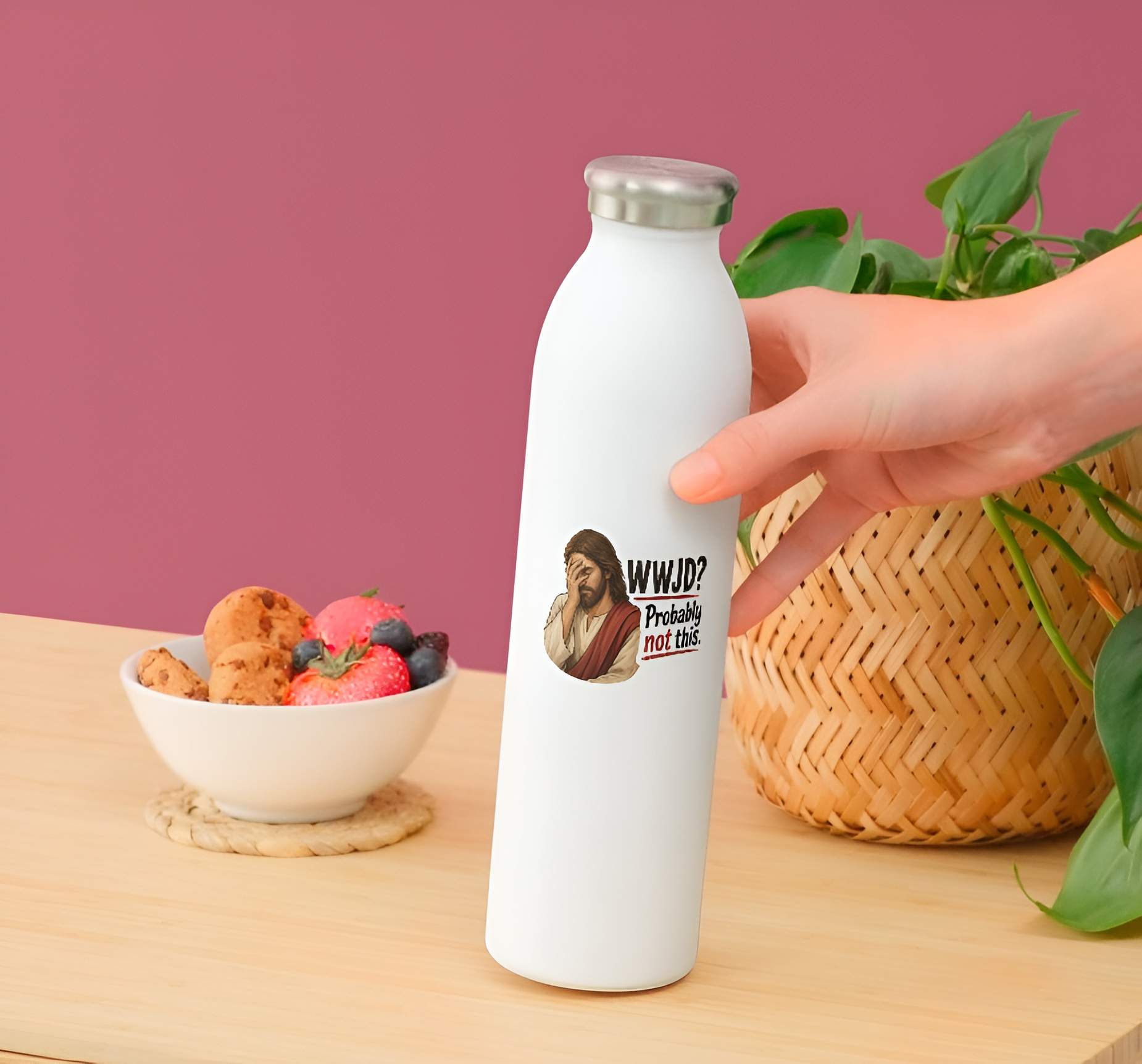 White water bottle with a Jesus wearing cream and red robes, holding his hand over his face in an exhausted facepalm gesture next to bold text reading "WWJD? Probably not this." design held by a hand on a wooden table with a bowl of snacks and a plant.