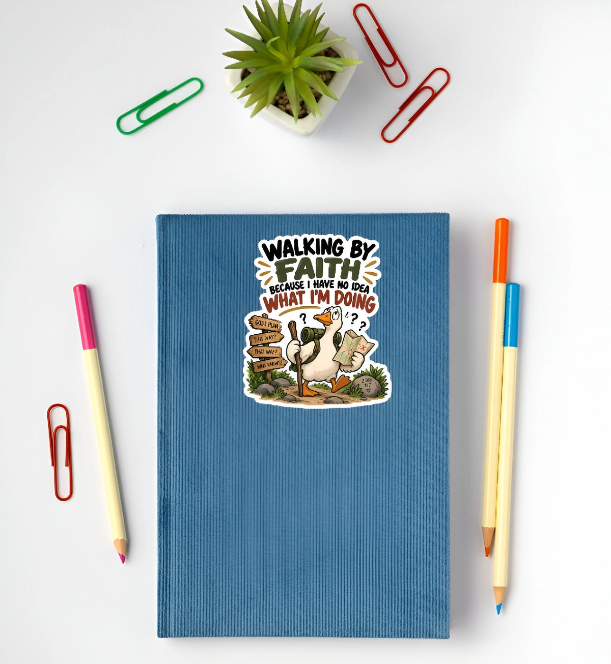 A blue hardcover notebook with a humorous cartoon graphic and the text 'WALKING BY FAITH BECAUSE I KNOW WHAT I'M DOING' lies on a white desk, surrounded by stationery props including paperclips, pencils, and a small potted succulent.