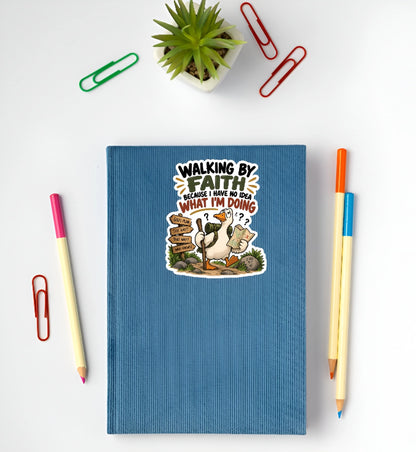 A blue hardcover notebook with a humorous cartoon graphic and the text 'WALKING BY FAITH BECAUSE I KNOW WHAT I'M DOING' lies on a white desk, surrounded by stationery props including paperclips, pencils, and a small potted succulent.