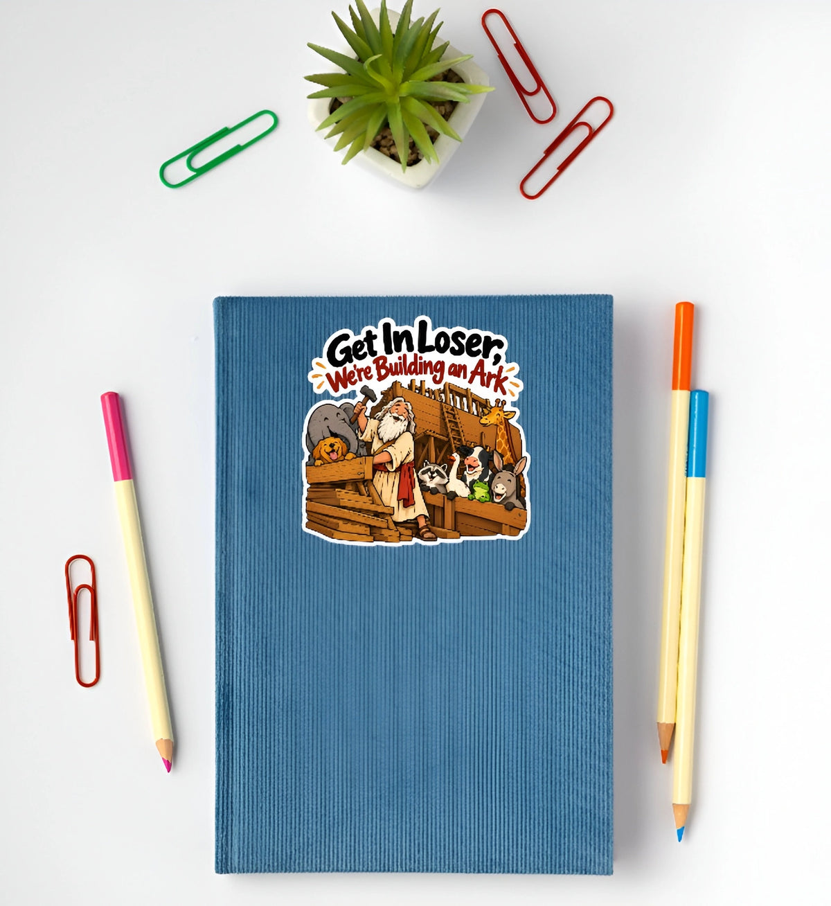 A blue ribbed hardcover notebook with a cartoon sticker on the cover reading 'Get In Loser We're Building an Ark', styled in a flat lay with colored pencils, paperclips, and a small potted succulent on a white background.