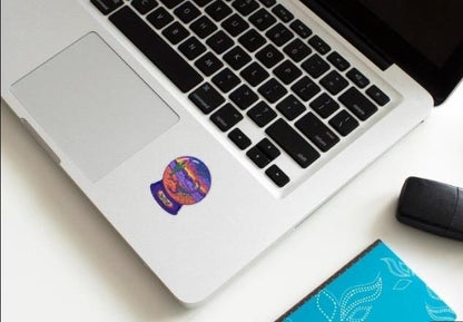 Laptop with a colorful sticker on the palm rest area, next to a mouse and notebook on a white surface.